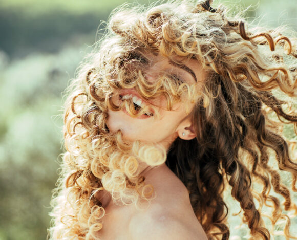 Mujer con rizos rubios al aire libre, sonriendo con el cabello en movimiento