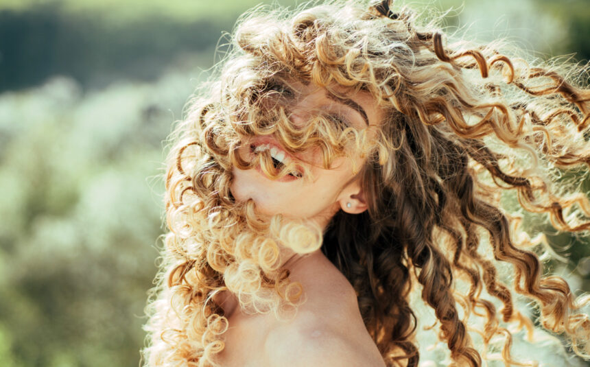 Mujer con rizos rubios al aire libre, sonriendo con el cabello en movimiento